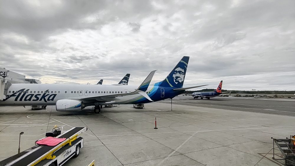 Several Alaska Airlines planes are parked at an airport gate under a cloudy sky, with a Hawaiian Airlines plane visible on the runway in the background. Ground service vehicles and equipment are nearby.