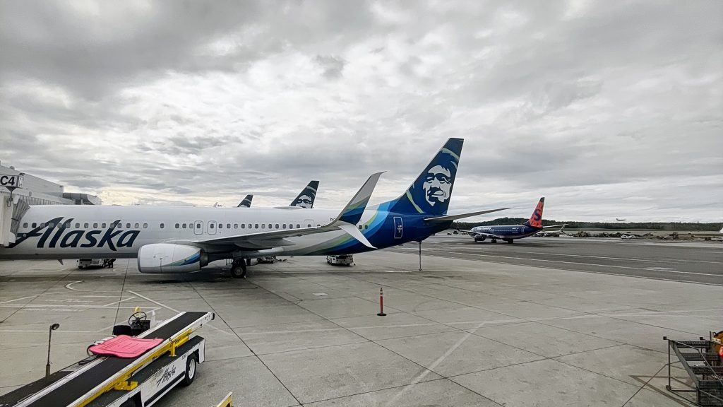 Several Alaska Airlines planes are parked at an airport gate under a cloudy sky, with a Hawaiian Airlines plane visible on the runway in the background. Ground service vehicles and equipment are nearby.