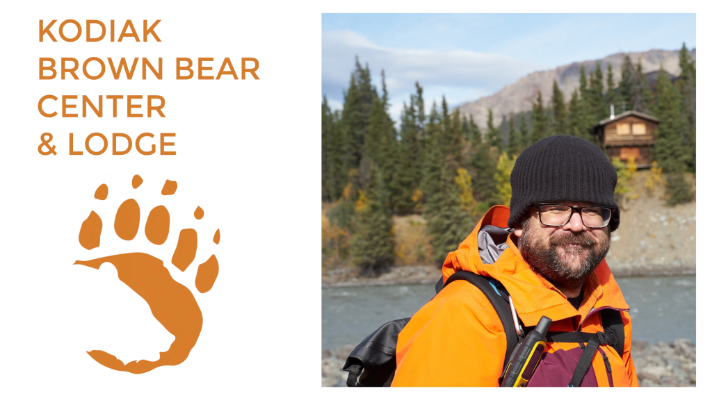 A man in an orange jacket and black beanie stands by a river with pine trees and a cabin in the background. Text reads “Kodiak Brown Bear Center Lodge” with a brown bear paw print illustration.