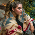A woman with light brown hair in a ponytail sits outdoors at Kodiak Lodge, draped in a patterned blanket with red, beige, and gray geometric designs. She looks to the side, surrounded by rocks and greenery.